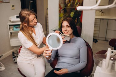Kapalı bir şekilde gülümseyen genç kadın diş minesi rengini stomatologla karşılaştır diş gölgesi ile dişçi koltuğunda otur iç mekan aydınlatma modern dolap sağlık hizmetleri diş minesi beyazlatma tedavisi.