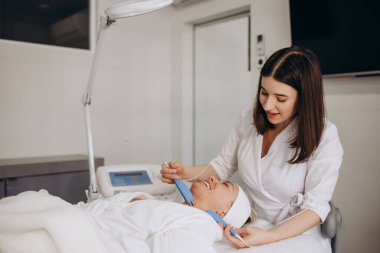 A doctor cosmetologist makes a microcurrent facial therapy to a young woman with a device in a beauty wellness salon.Cosmetology and professional skin care.