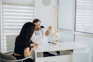 Kadın hasta, doktorun muayenehanesinde sırtıyla ilgili sorunları hakkında doktorla konuşuyor. Doktor ve hasta koruyucu maske takıyor. Doktora odaklan..