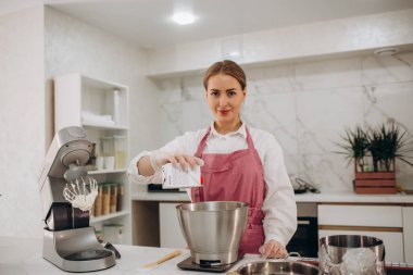 El çırpıcı ve krema tutar, diğeri süt döker. Beyaz mutfak. Dondurma, dondurma. Yan görüş. Masanın üzerinde havlular, silikon kalıplar, çikolata, pasta şırıngası, şekerleme, waffle var..