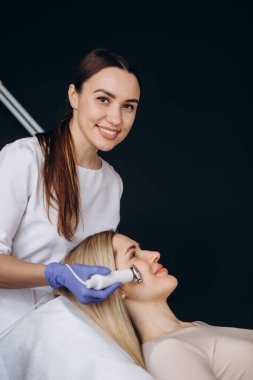 Güzellik uzmanı kozmetik uzmanı, mikro akım terapi cihazıyla yüzünden bir işlem yapıyor. Klinikte müşteri için donanım kozmetolojisi.