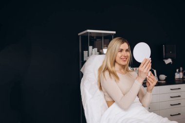 Güzel ve mutlu genç bir kadın tıbbi sandalyede oturup aynaya bakıyor. Hyaluronik asit doldurucu ya da botulinyum toksin iğneleriyle başarılı bir güzellik tedavisinden sonra tatmin oluyor.
