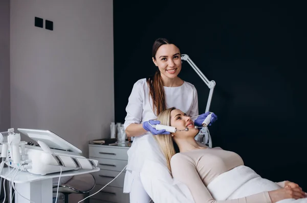 stock image Beautiful relaxed woman receiving apparatus facial microcurrent treatment from therapist in beauty wellness salon, side view. Electrical impulses in cosmetology concept