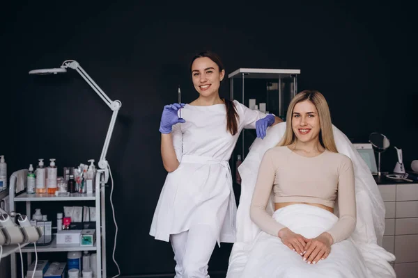 Elinde şırıngayla bir kozmetik uzmanının portresi ve kozmetoloji kliniğinde kanepede oturan bir kadın müşteri..