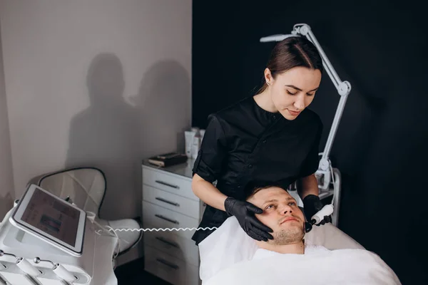 Cosmetologist is cleaning the young man\'s face from acne and scars by ultrasonics
