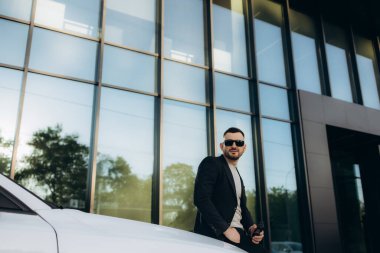 Yakışıklı, gülümseyen, mutlu, sakallı adam cep telefonu arıyor ve modern ofis merkezinin yakınındaki caddedeki yeni beyaz arabasına gidiyor.