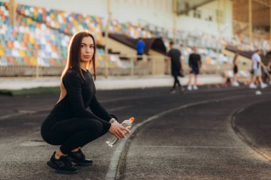 Sporcu kadın mavi pistte oturuyor ve esniyor. Kadın koşucu, ayakkabısına dokunarak bacak kaslarını esnetiyor. Isınan bayan koşucu, koşu pistinde oturarak esneme egzersizleri yapıyor..