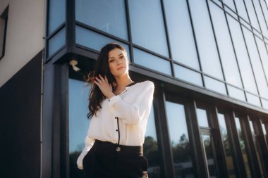 Beyaz bluzlu bir iş kadını, elinde akıllı bir telefonla sokakta, bir iş merkezinin arka planında. İnternette çalışıyorum. Yüzünde ciddi bir ifade