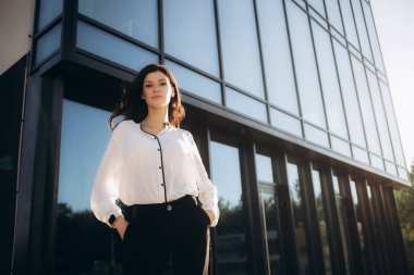 Beyaz bluzlu bir iş kadını, elinde akıllı bir telefonla sokakta, bir iş merkezinin arka planında. İnternette çalışıyorum. Yüzünde ciddi bir ifade