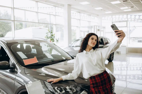 Happy woman customer buyer client in white shirt do selfie shot mobile cell phone show ok gesture choose auto want buy new automobile in car showroom vehicle salon dealership store motor show indoor.