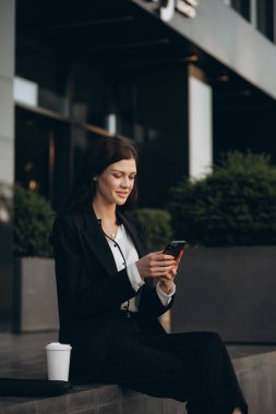 Beyaz iş kadını yürüyor, akıllı telefon kullanıyor ve sokakta kahve içiyor. Kahve molası sırasında ofis binasının yakınında mesaj yazan çekici bir kadın..