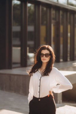 Kıvırcık saçlı, kendine güvenen zarif bir genç kadın, resmi olarak giyinmiş, iş merkezinin arka planında duruyor, elinde belgelerle bir dosya tutuyor, kameraya bakıyor, gülümsüyor.