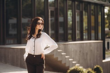 Kıvırcık saçlı, kendine güvenen zarif bir genç kadın, resmi olarak giyinmiş, iş merkezinin arka planında duruyor, elinde belgelerle bir dosya tutuyor, kameraya bakıyor, gülümsüyor.