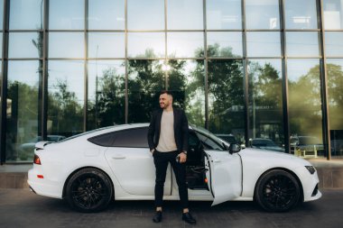 Yakışıklı, gülümseyen, mutlu, sakallı adam cep telefonu arıyor ve modern ofis merkezinin yakınındaki caddedeki yeni beyaz arabasına gidiyor..