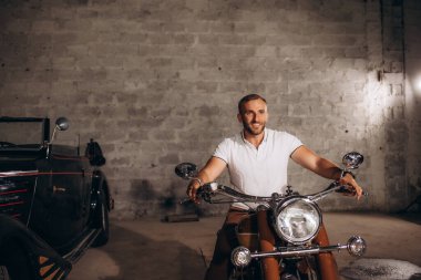 Bir adam garajda motosikletin üstünde oturuyor..