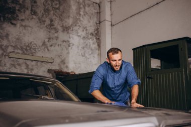 Bir adam garajdaki eski bir arabanın tozunu alıyor..