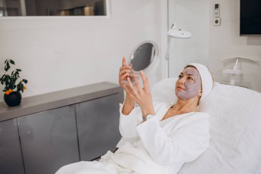 Güzel ve mutlu genç bir kadın tıbbi sandalyede oturup aynaya bakıyor. Hyaluronik asit dolguları ya da botulinyum toksin enjeksiyonlarıyla başarılı bir güzellik tedavisinden sonra tatmin oluyor. Yüksek kalite fotoğraf