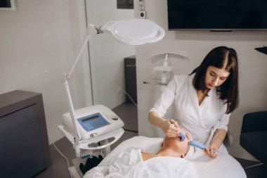 Bir doktor kozmetik uzmanı güzellik salonunda cihazı olan genç bir kadına mikro akım yüz terapisi yapıyor. Kozmetoloji ve profesyonel cilt bakımı. Yüksek kalite fotoğraf