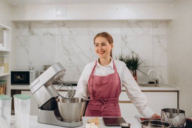 Pasta şefi şekerlemeci genç beyaz kadın mutfakta. Önünde tezgahta bir pasta var. O, bir tabak üzerinde bir parça kek tutar. Tatlı keklerin yanında, şeker kasesinde bir fincan şeker.. 