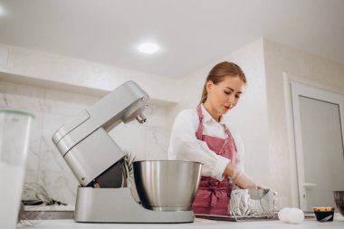 Oldukça neşeli genç bir kadın, beyaz üniformalı ve şapkalı profesyonel bir şekerci, yumurta beyazlarını çırpmak için elektrikli gezegen mikseri kullanıyor, kaseye şeker döküyor.. 