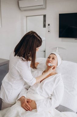 Yetenekli kozmetikçi müşteri için sağlıklı yüz maskesi yapıyor. Ayakta duruyor ve dişinin çenesine dokunuyor. Genç kadın gözleri kapalı uzanıyor ve dinleniyor. Yüksek kalite fotoğraf