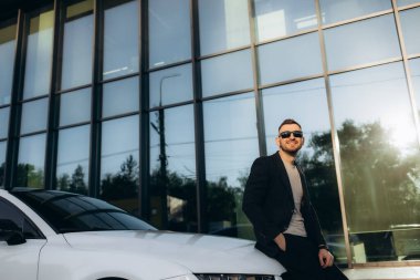 Yakışıklı, gülümseyen, mutlu, takım elbiseli sakallı adam cep telefonu arıyor ve modern ofis merkezinin yakınındaki caddedeki yeni beyaz arabasına gidiyor. Yüksek kalite fotoğraf