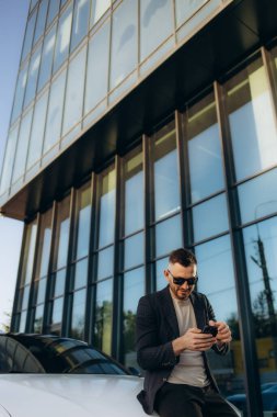 Yakışıklı, gülümseyen, mutlu, takım elbiseli sakallı adam cep telefonu arıyor ve modern ofis merkezinin yakınındaki caddedeki yeni beyaz arabasına gidiyor. Yüksek kalite fotoğraf
