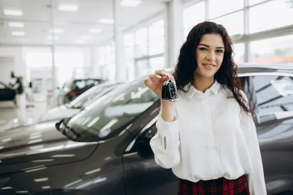 Happy brunette woman buying new cute car, raising hand up with automatic key and smiling, showroom interior, copy space. Pretty curly lady customer purchasing yellow female auto, getting in. High quality photo