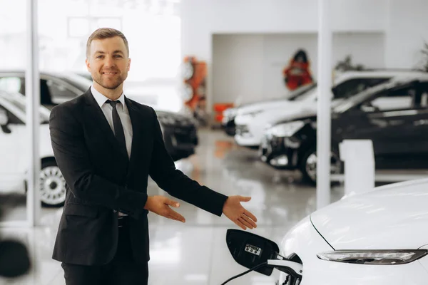 Portrait of a confident sales manager in the suit near the car charging station, selling electric cars in the showroom. High quality photo