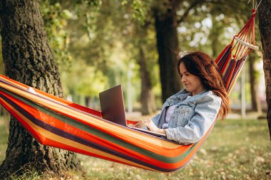 Bilgisayarda internette gezinen güzel genç bir kadın hamakta yatarken, arka planda pitoresk bir tropikal park. 