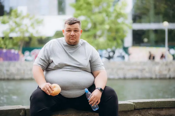 Stadyumda antrenmanda hamburger yiyip su içen şişman bir adam..