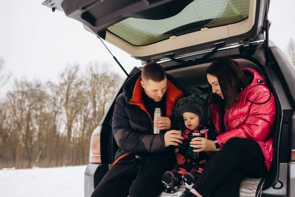 Happy family with dog on vacation during the winter holidays near road. Dressed in warm clothes sitting on the trunk of a car and drinking tea from a thermos. space for text. Winter vacation. High quality photo