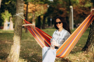 Bir kadın hamakta otururken su içer.