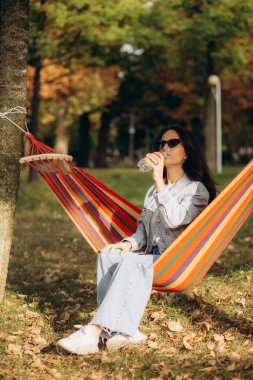 Bir kadın sonbahar parkında hamakta dinlenirken su içer.