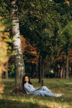 Görüntülü görüşme için dizüstü bilgisayar ve kablosuz kulaklık kullanırken yaz gün boyunca bağdaş ile Park yeşil çimenlerin üzerinde oturan genç ve güzel kadın portresi. 