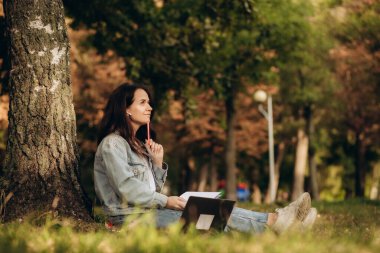 Öğrenci dışarıda çalışıyor. Modern bir kızın ağaçların altında çimlerde oturması, dizüstü bilgisayar, kahve, defter, bardaklar ve parktaki akıllı telefonla. Eller klavyede. Ekran modeli. Mesaj için yer var. Yüksek kalite fotoğraf