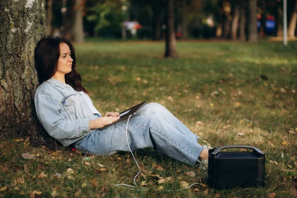 A woman works on a laptop under a tree and charges it from a portable charging station. High quality photo