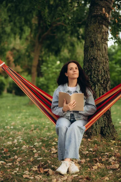 a woman reads a book in the park while sitting in a hammock