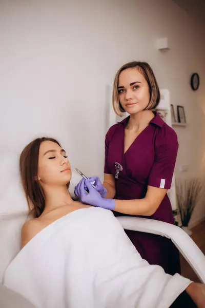Professional female beautician makes a rejuvenating injection to a young woman who lies relaxed with her eyes closed in a modern beauty salon. Concept of smoothing wrinkles with injections. High quality photo