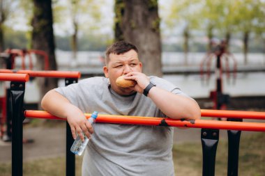 Şişman bir adam parkta oyun alanında hamburger yiyor.