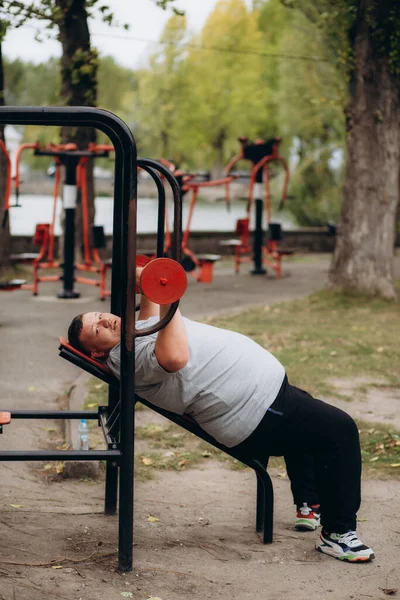 Şişman bir adam parkta egzersiz makinesinde halter basıyor.