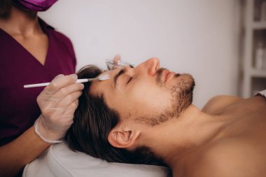 Kadın güzellik uzmanı yüzüne kil maske takarken, gözleri kapalı, başını yumuşak havluya sarmış genç bir adamın yan görüntüsü. Yüksek kalite fotoğraf