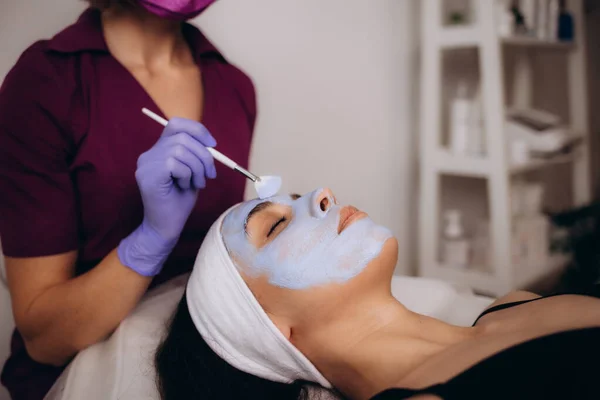 Cosmetologist applying blue mask to face woman client, rejuvenation procedure to beautiful in a beauty salon. High quality photo