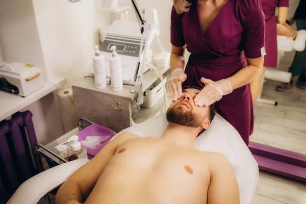 Beautician applying scrub onto young man's face in spa salon. High quality photo