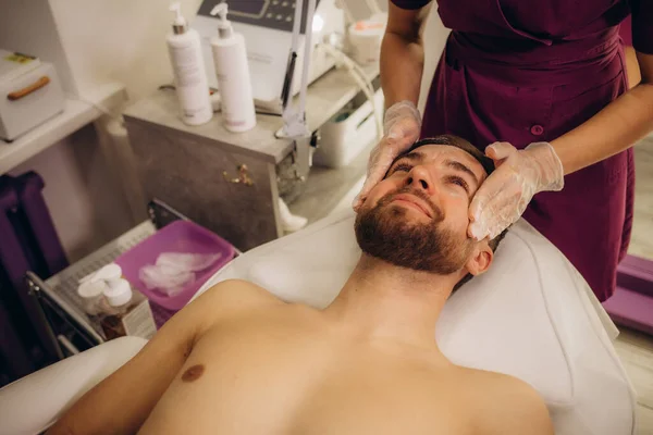 Beautician applying scrub onto young man\'s face in spa salon. High quality photo