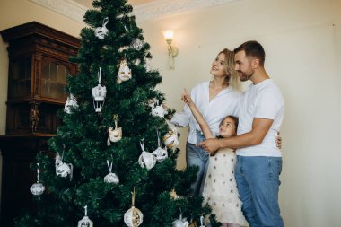 Baba, anne ve kızı Noel ağacı süsler. Mutlu sevgi dolu aile hediyeleri. Altında hediyeler olan Noel ağacı. dekore edilmiş oturma odası.