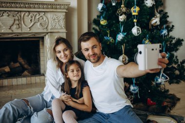 Mutlu Noeller ve mutlu Noeller. Genç bir aile bir Noel ağacının arka planında selfie çekiyor.