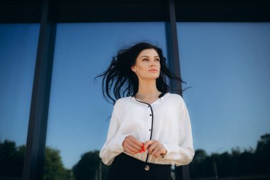 Kıvırcık saçlı, kendine güvenen zarif bir genç kadın, resmi olarak giyinmiş, iş merkezinin arka planında duruyor, elinde belgelerle bir klasör tutuyor..