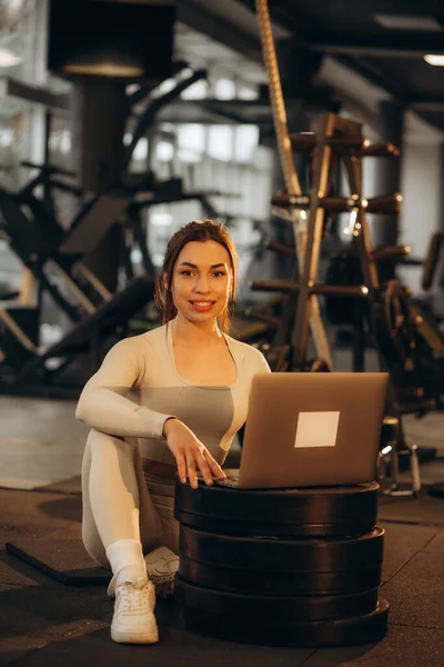 Smiling girl with laptop in a gym. Athlete with dumbbells on a blurred background. Active lifestyle concept.