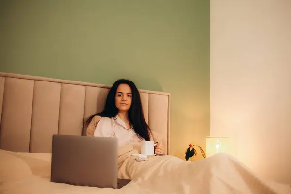 Upset sick lady in pajamas holding cup of hot drink while sitting on bed in front of modern notebook. High quality photo